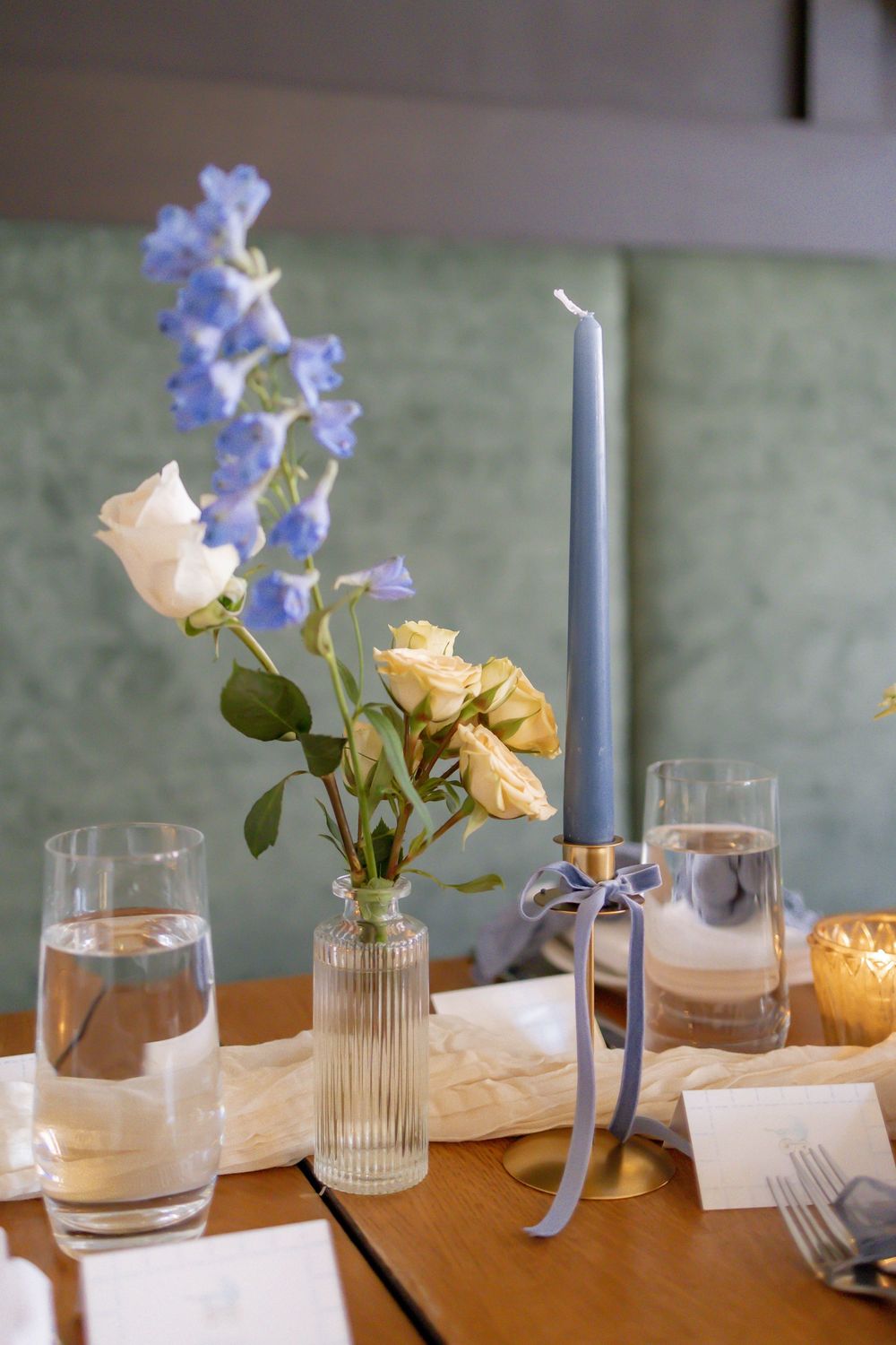Elegant baby shower table setting with blue taper candle, white and yellow roses, and blue delphinium in glass vases.