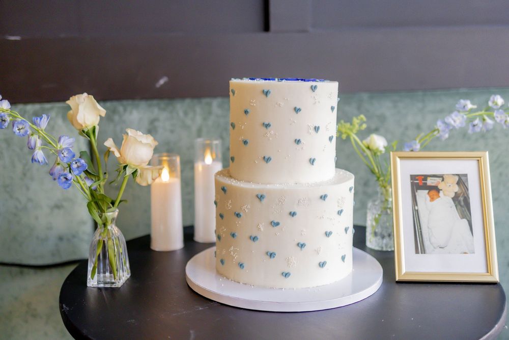 Two-tier white wedding cake with blue hearts, flanked by candles, white roses, and a framed wedding photo.