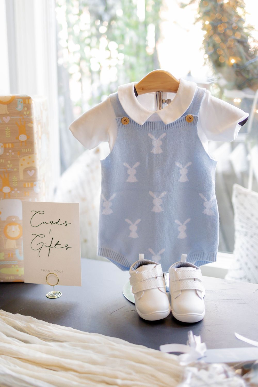 Blue bunny knit baby romper displayed on hanger with white shoes and Cards & Gifts sign at baby shower.