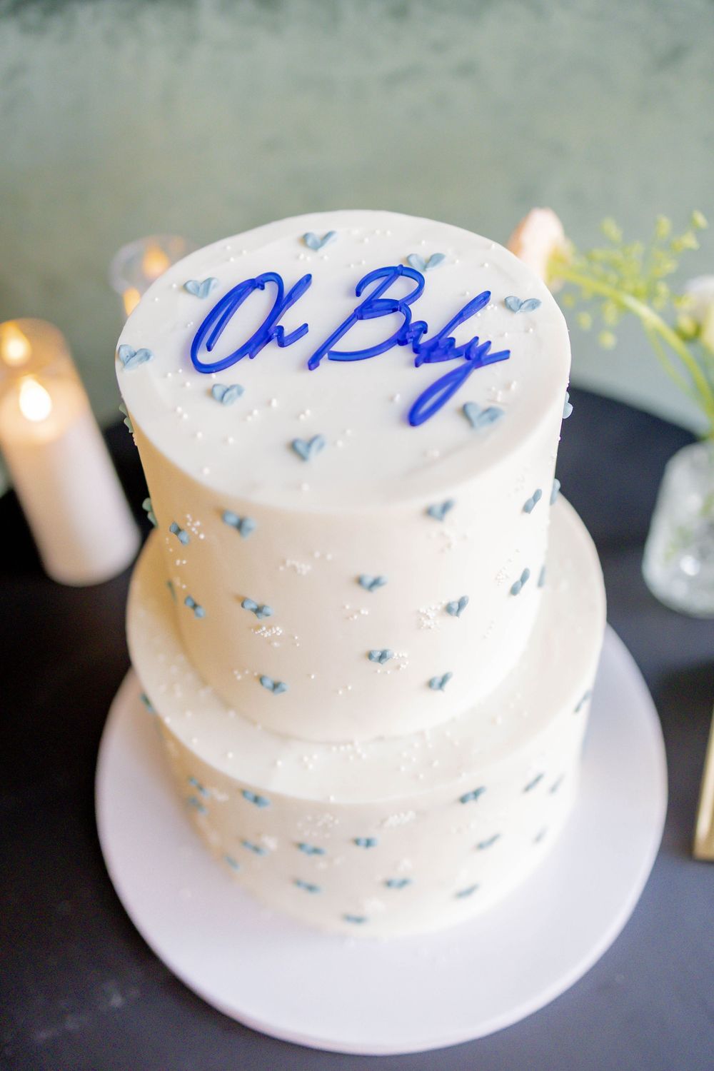 Two-tier white baby shower cake with blue heart details and bold blue Oh Baby topper on white cake stand.