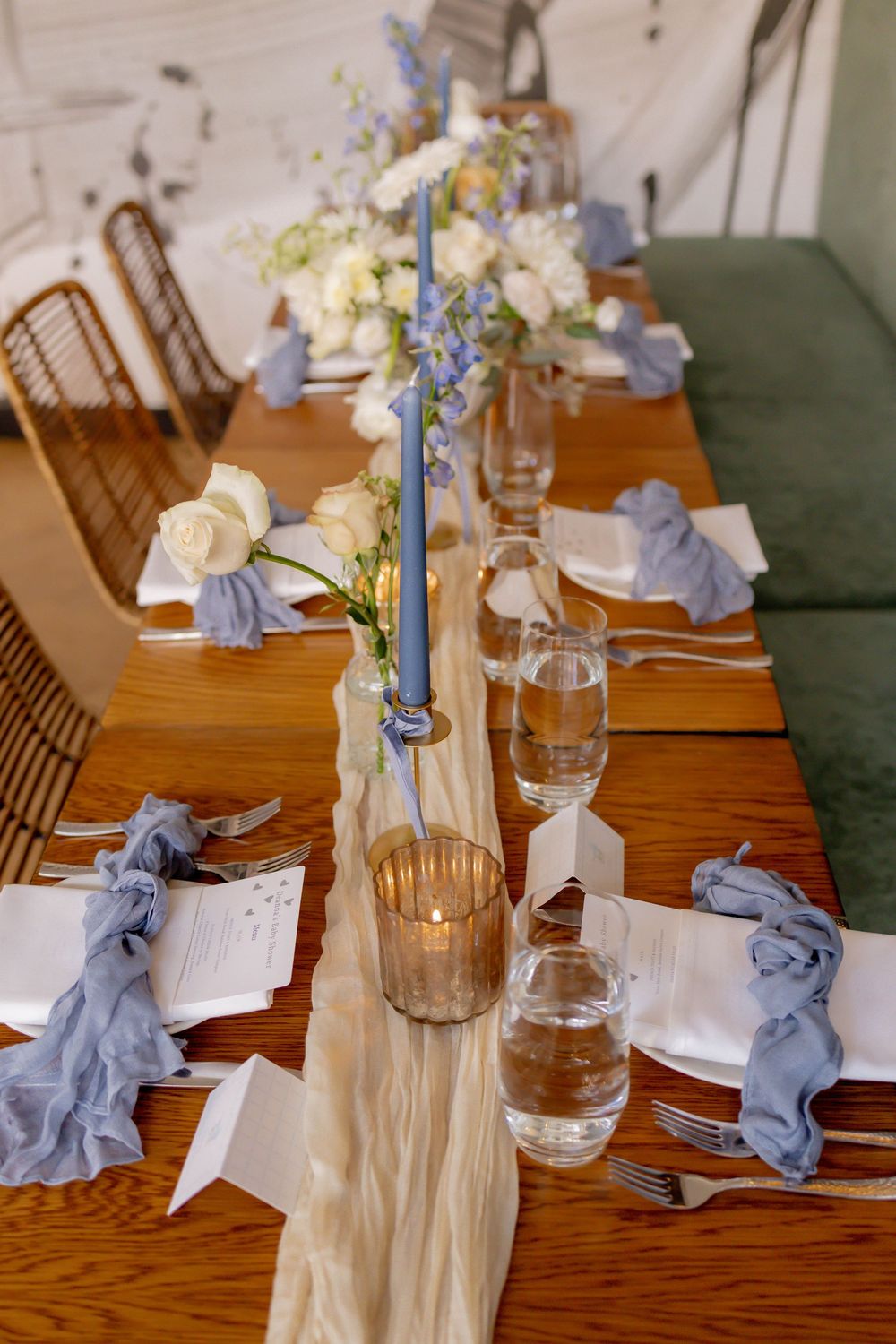 Elegant wedding reception table with blue candles, white roses, dusty blue napkins, and a cream table runner on a wooden table.