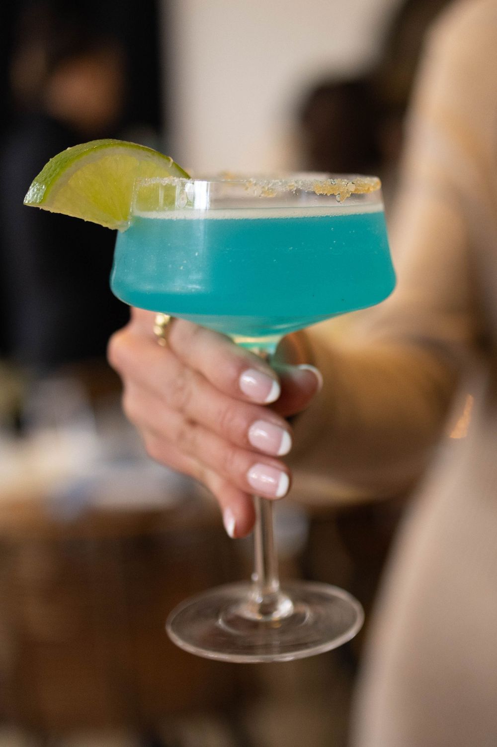 Woman holding a blue margarita cocktail with a salted rim and lime garnish in a classic margarita glass.