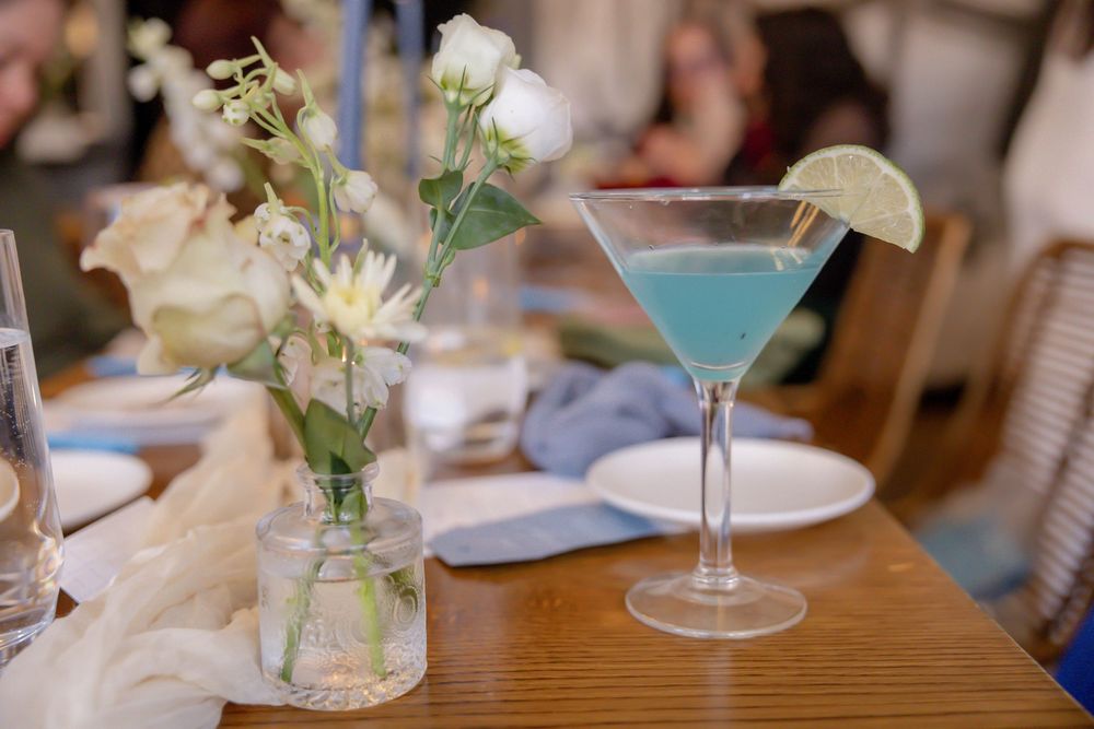 Blue martini cocktail with lime garnish beside white floral arrangement on wooden table.