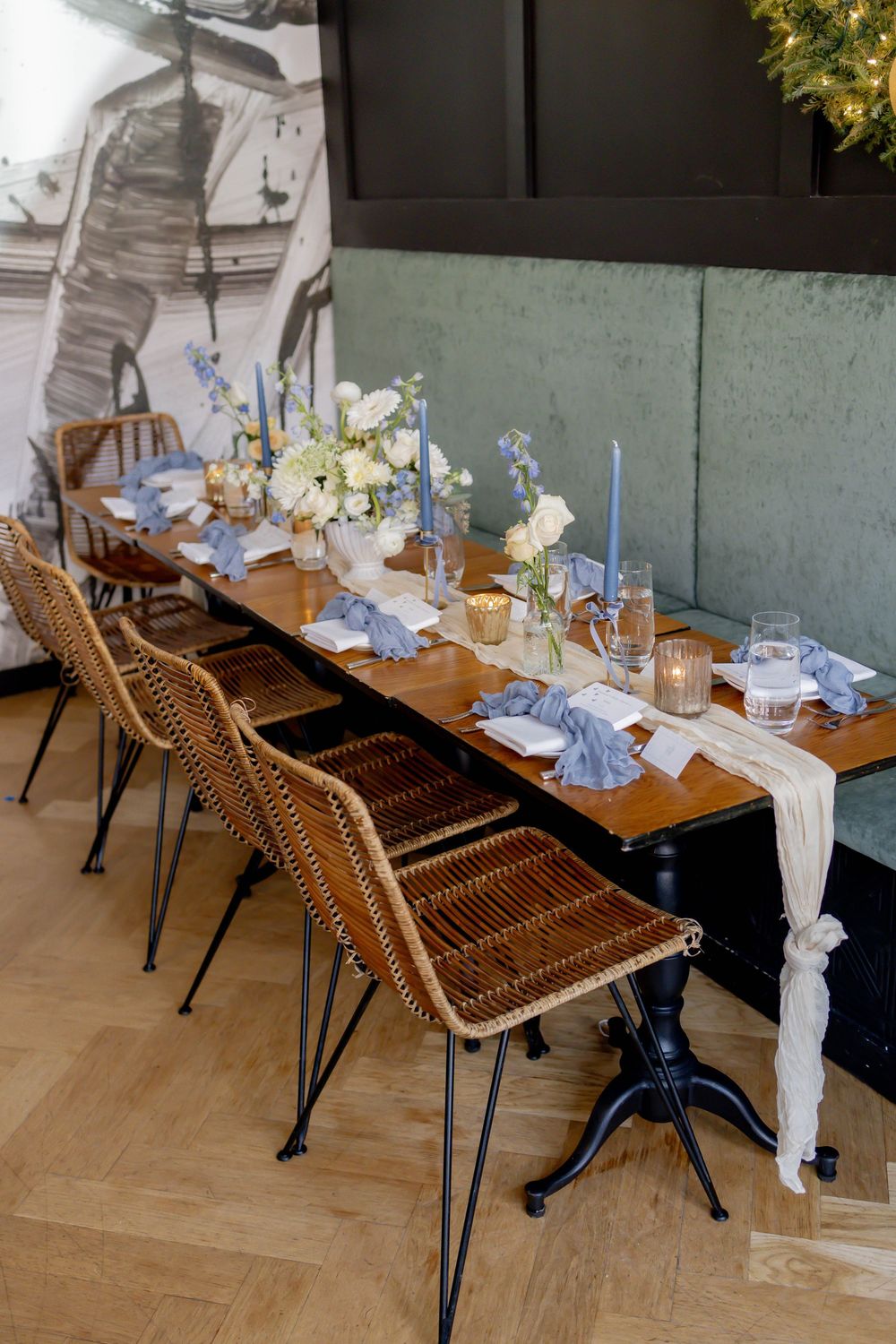 Elegant dining table with wicker chairs, blue taper candles, white floral centerpieces, and blue linen napkins on herringbone wood floors.