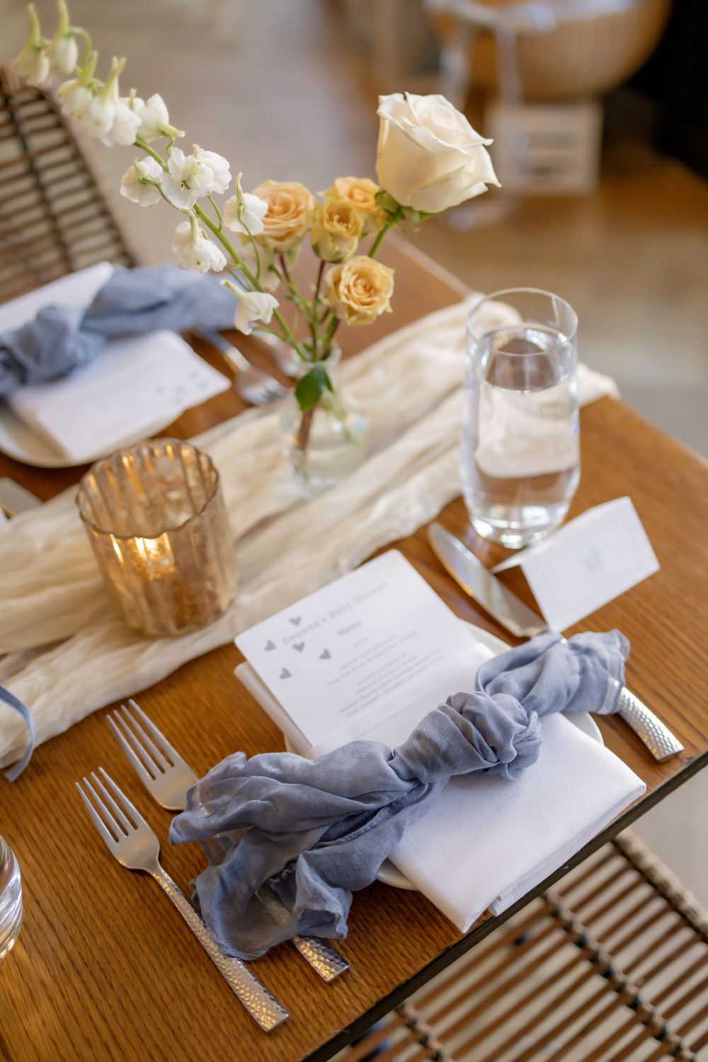 Elegant wedding table setting with blue linen napkins, white menu cards, gold candles, and peach roses in a glass vase.