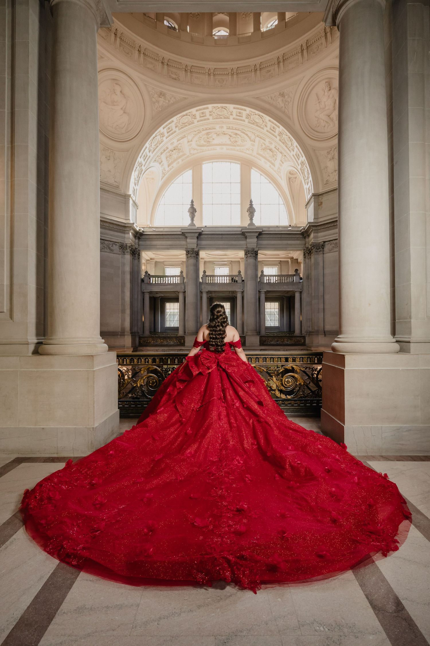 Woman in blue ballgown on ornate gold staircase in elegant mansion with arched windows and chandelier.