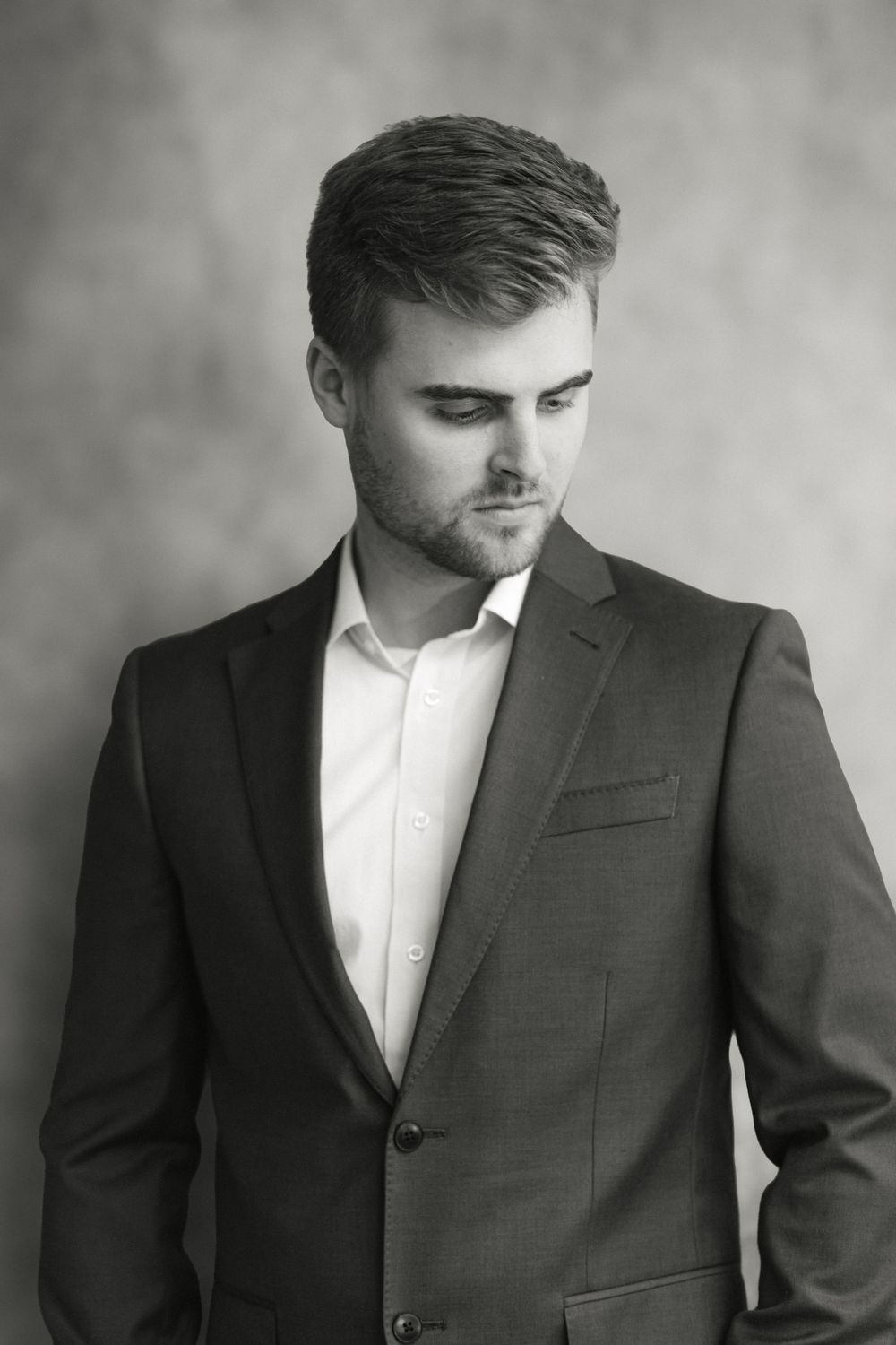 Pensive young man in dark suit looks downward in moody black and white studio portrait.