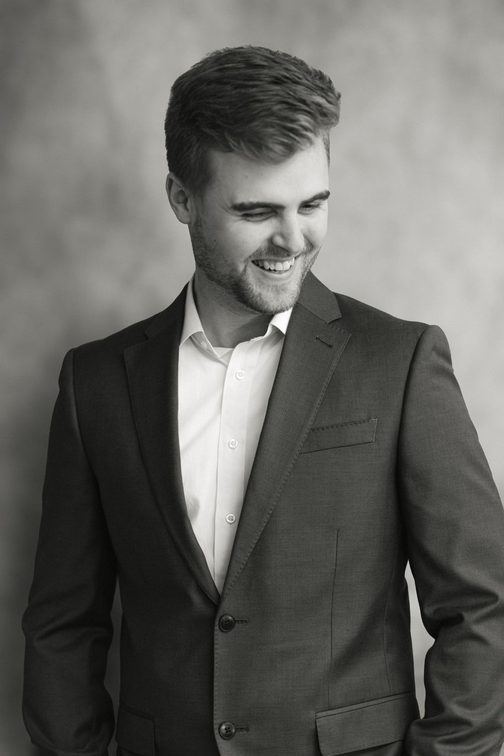 Smiling young man in dark suit laughs candidly in stylish black and white studio portrait.