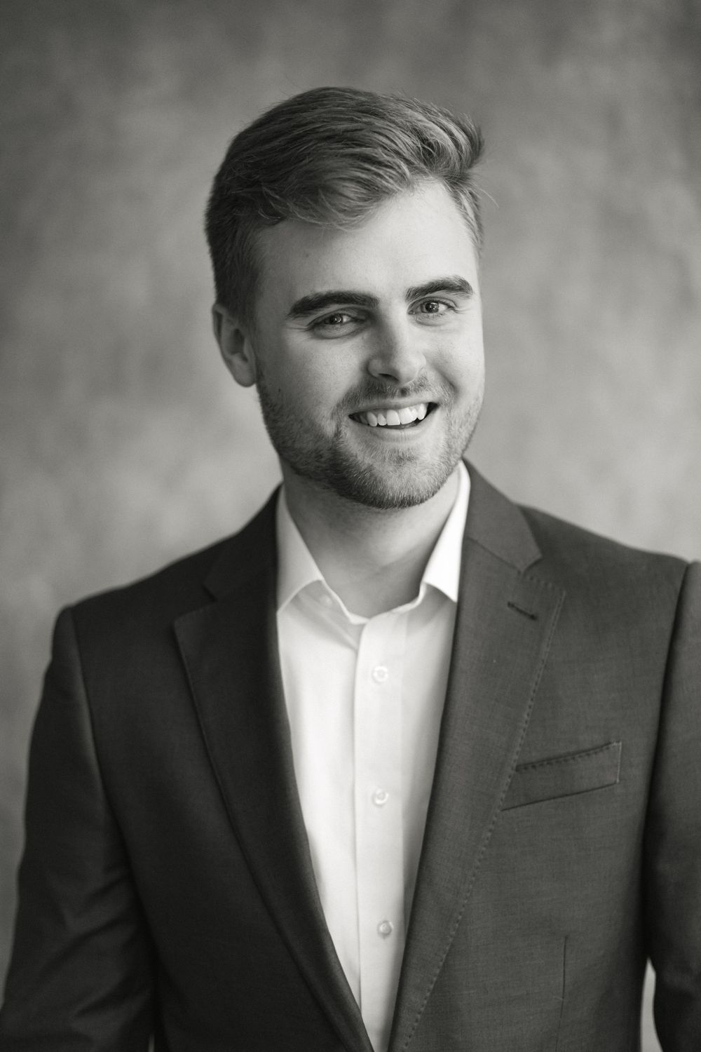 Smiling young man with light hair wearing a dark blazer and white shirt in a black and white professional portrait.