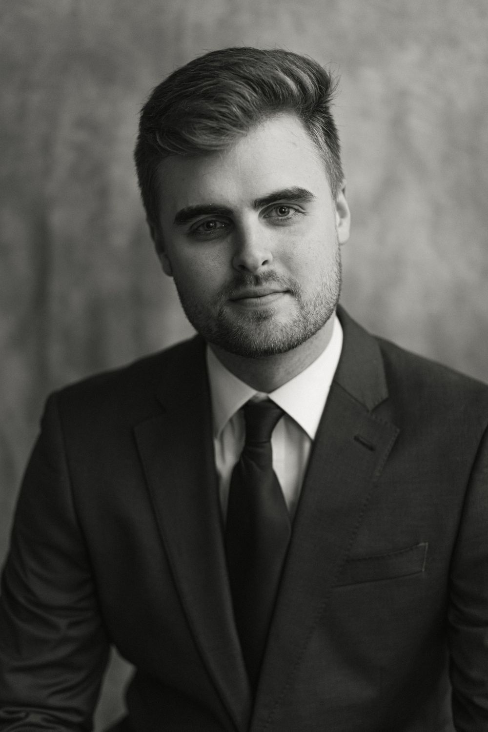 Serious young man in black suit and tie poses in classic black and white studio portrait.