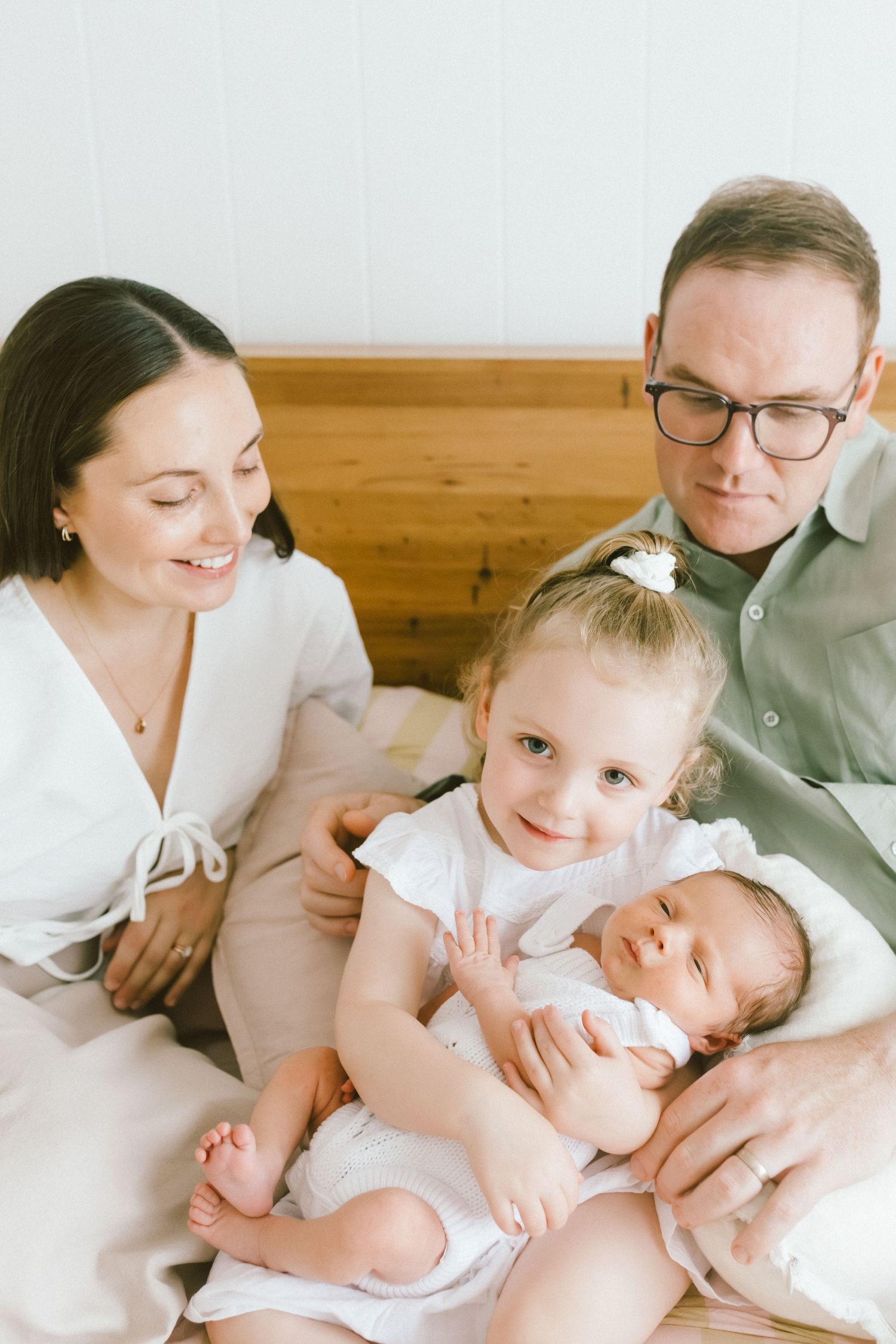 A happy family of four with parents, toddler girl, and newborn baby sitting together on a bed.
