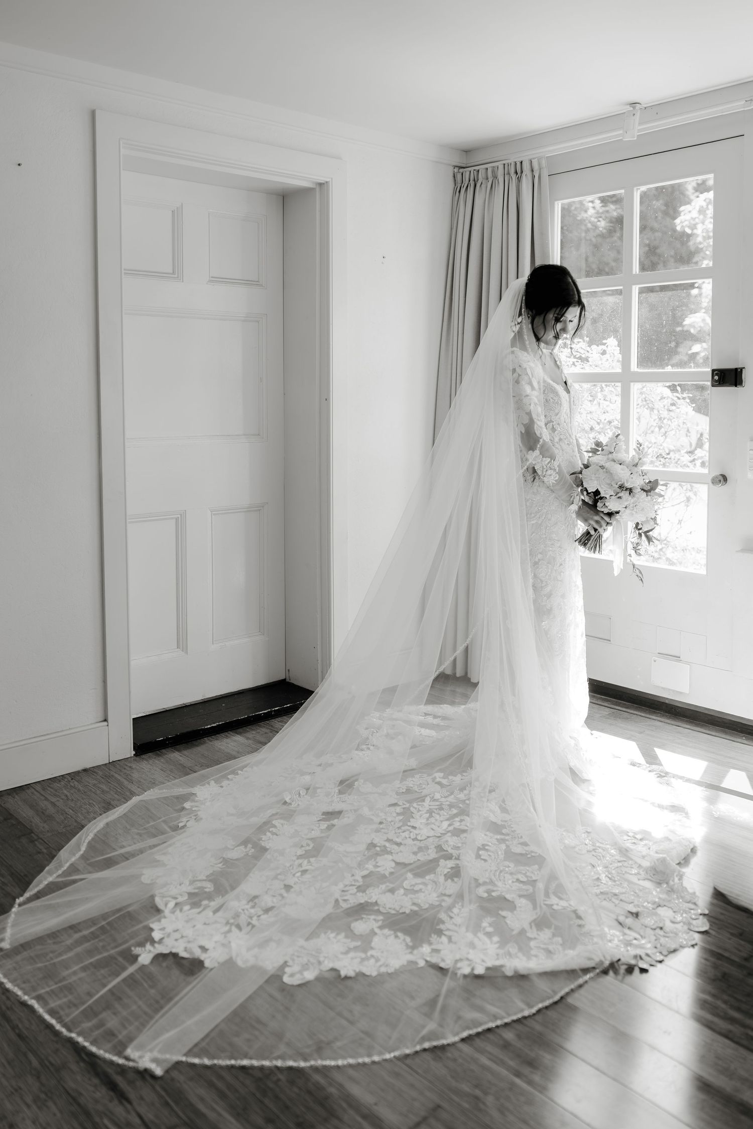Bride in lace wedding gown with cathedral veil stands by window holding bouquet, black and white photo.