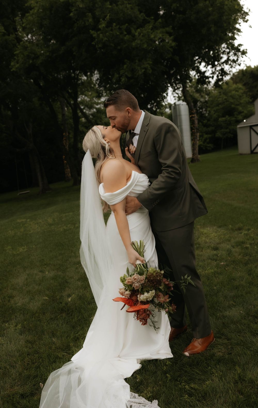 Bride and groom share romantic kiss outdoors, bride holding lush red and white wedding bouquet with veil.