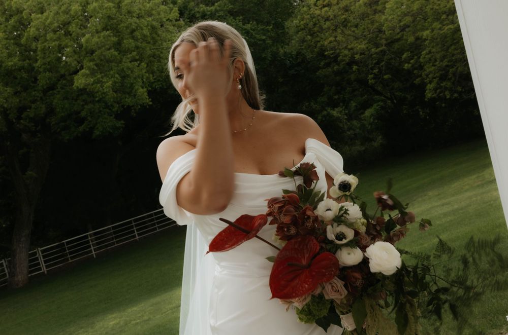 Emotional bride in white off-shoulder gown holding red and white bouquet outdoors on lush green lawn.