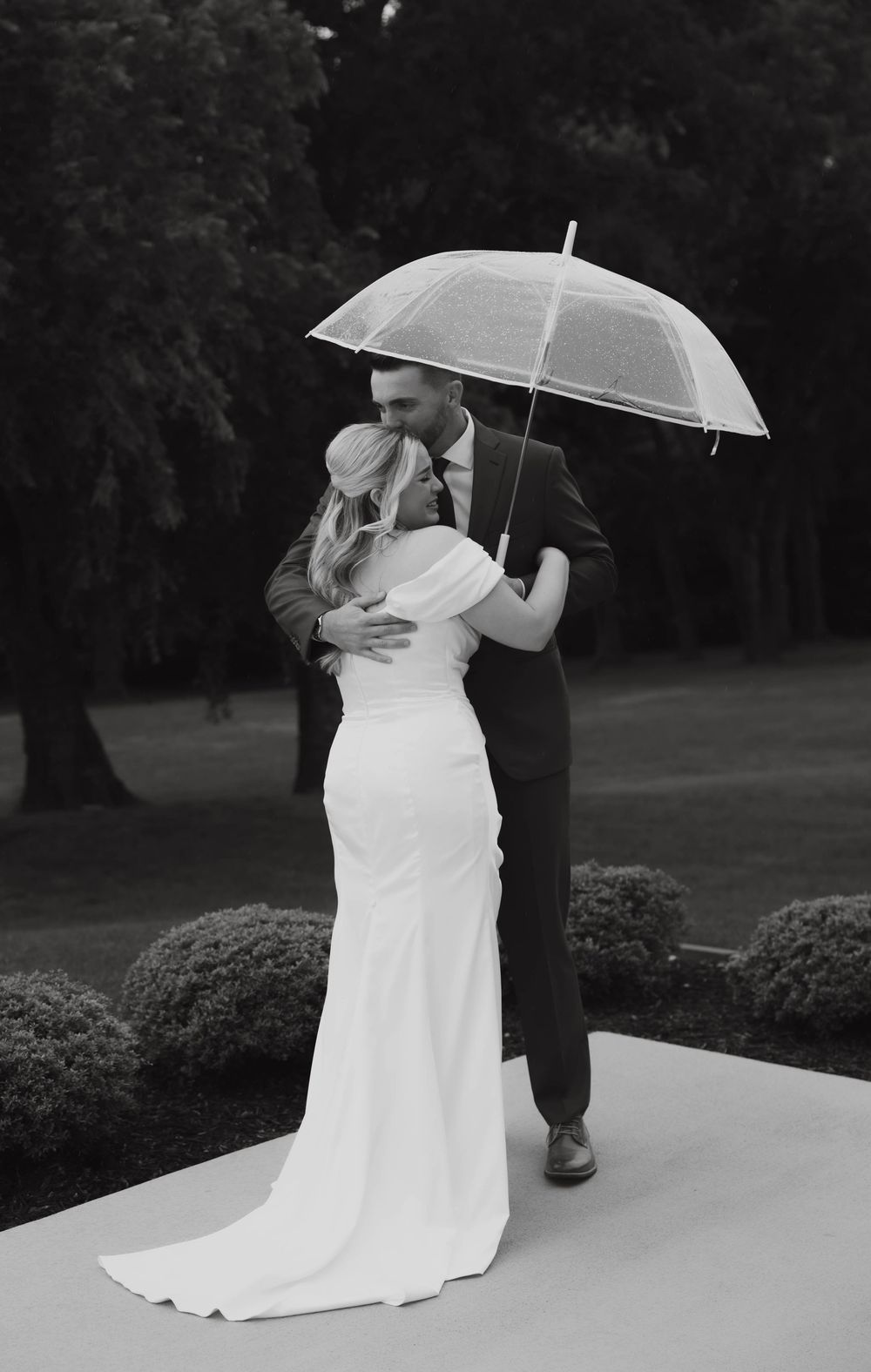 Black and white photo of bride and groom kissing under clear umbrella on garden pathway.