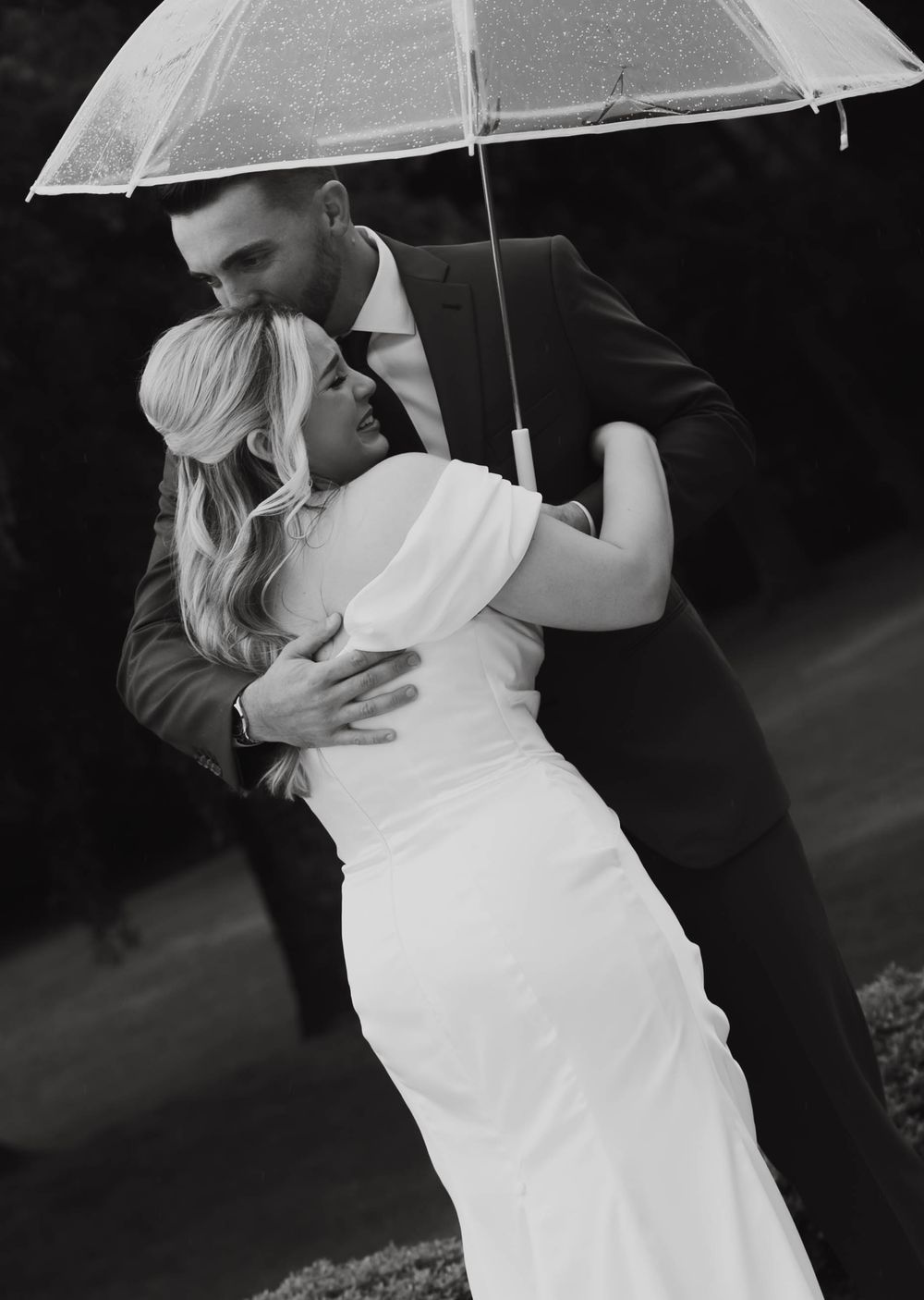 Black and white romantic photo of bride and groom embracing under clear umbrella in the rain.