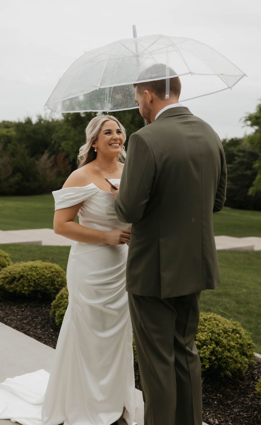 Smiling bride in white off-shoulder gown shares first look with groom in olive suit under umbrella.