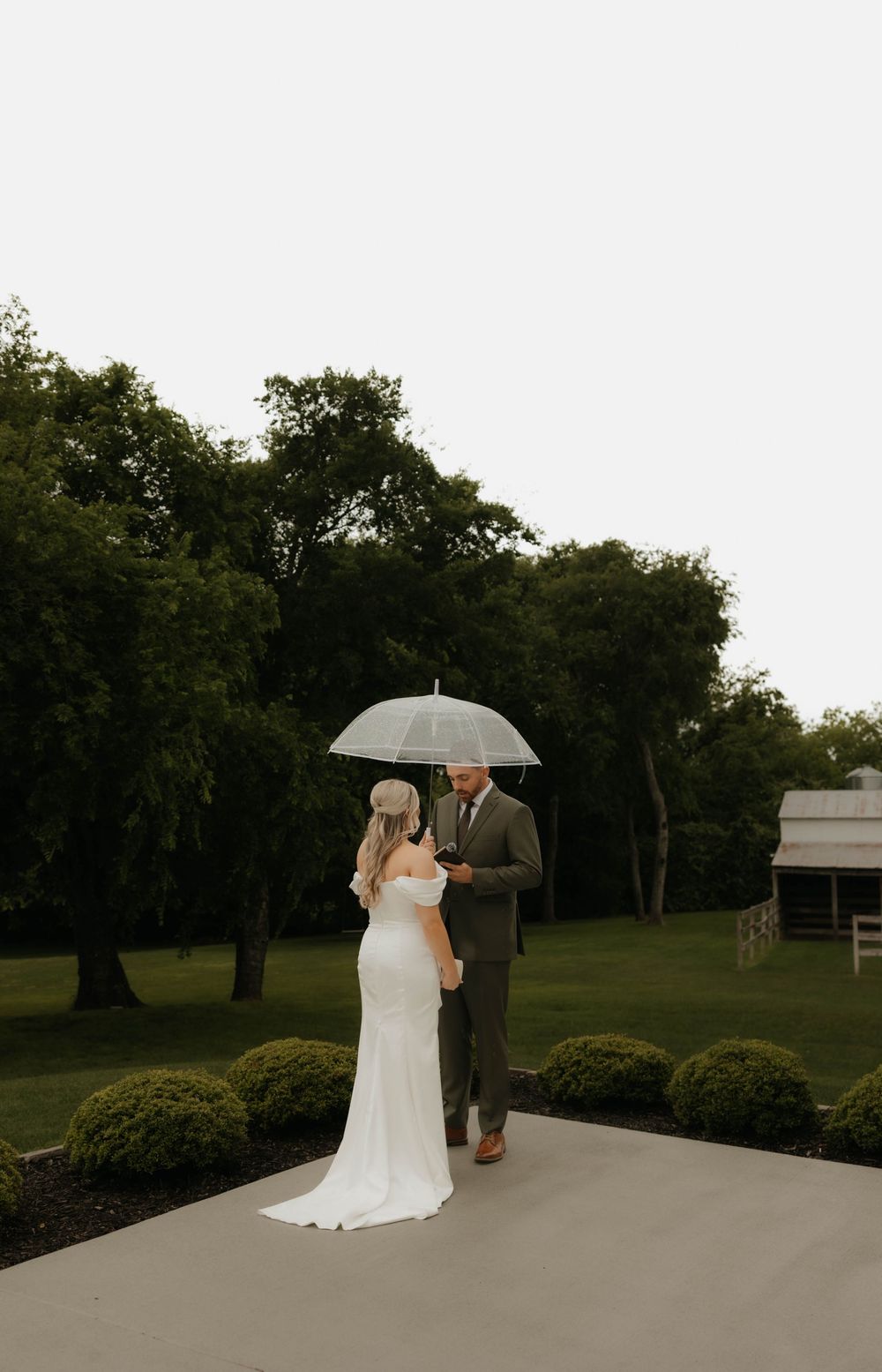 Bride and groom share intimate moment under clear umbrella on elegant garden pathway on rainy day.