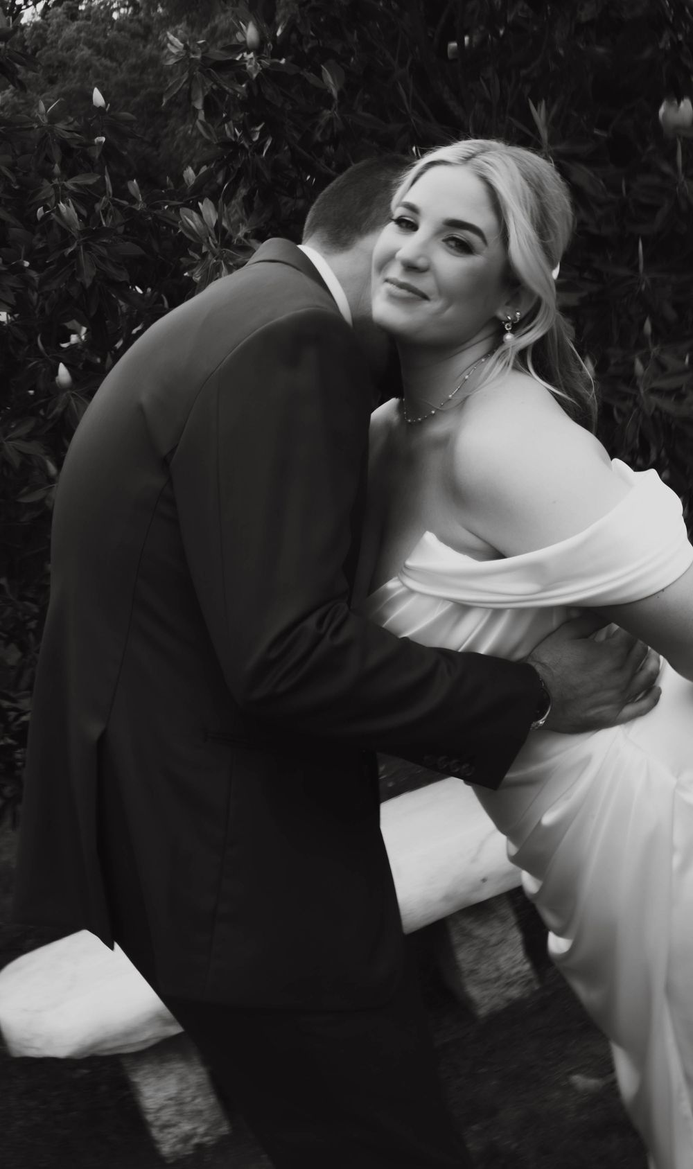 Black and white photo of smiling bride in off-shoulder dress embracing groom at wedding.
