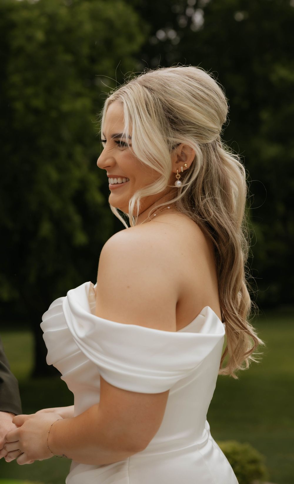 Smiling bride in an off-shoulder white wedding gown with gold pearl earrings outdoors.