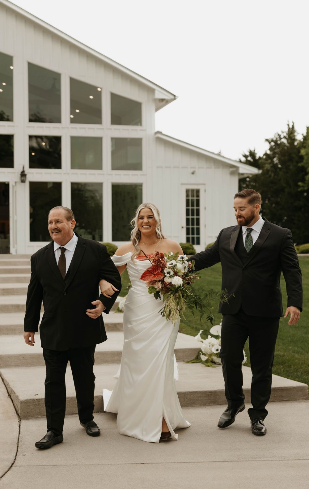 Smiling bride in white off-shoulder gown with red bouquet walks with two men outside white barn venue.