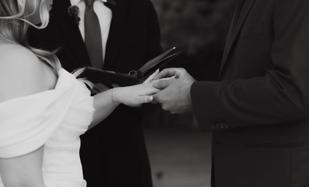 Black and white close-up of officiant placing wedding ring on brides hand during ceremony.