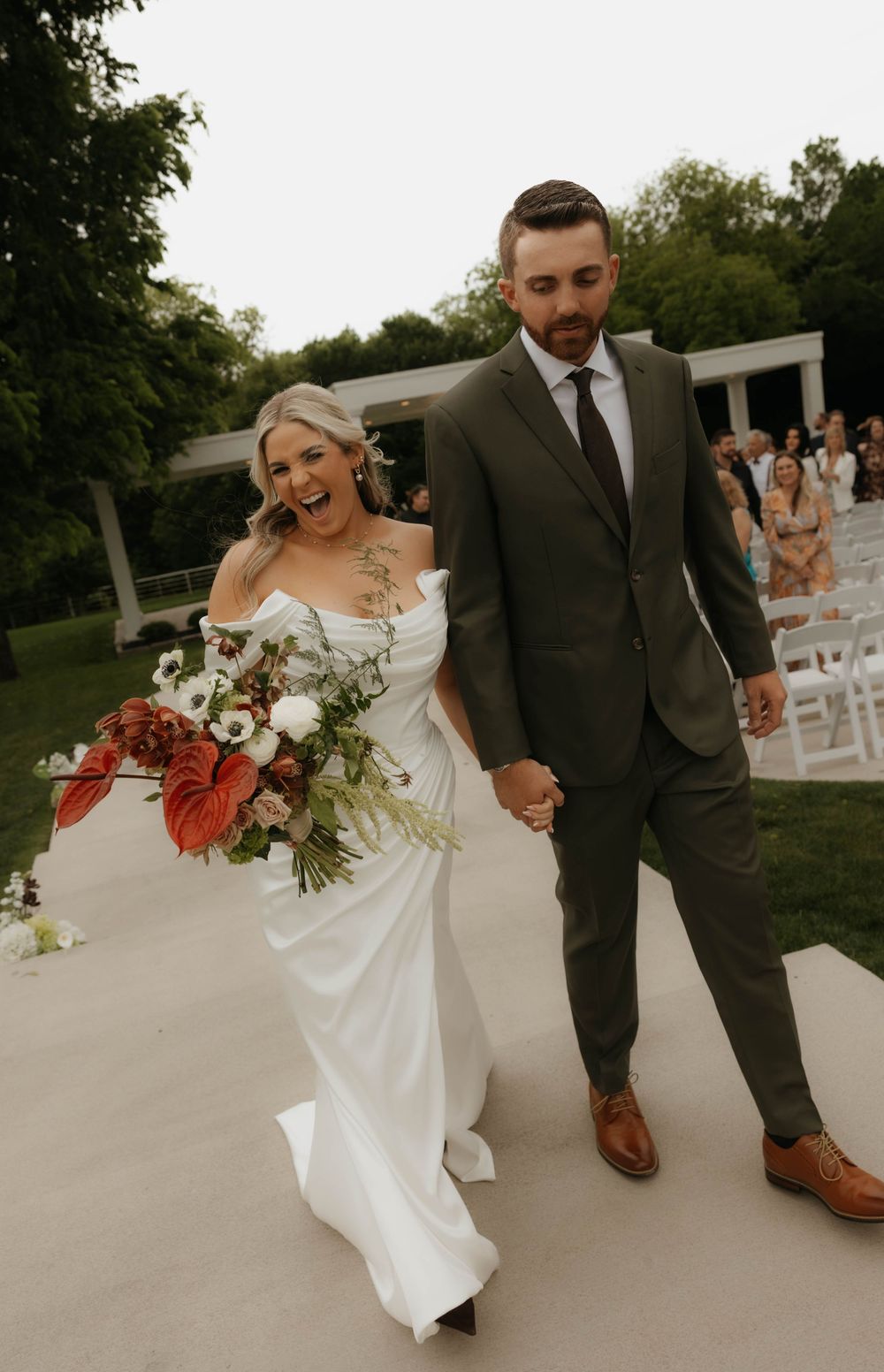 Joyful bride and groom walk back down aisle after wedding ceremony holding hands with large floral bouquet.