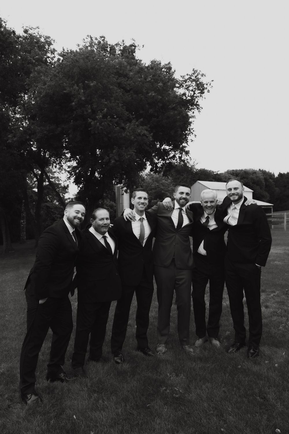 Black and white photo of groom and groomsmen laughing together outdoors at a wedding celebration.