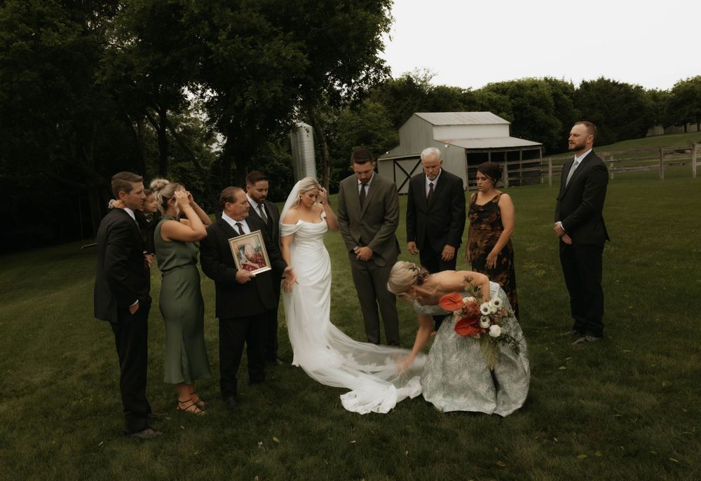 Wedding party gathered outdoors with bride in white gown holding bouquet near rustic barn venue.