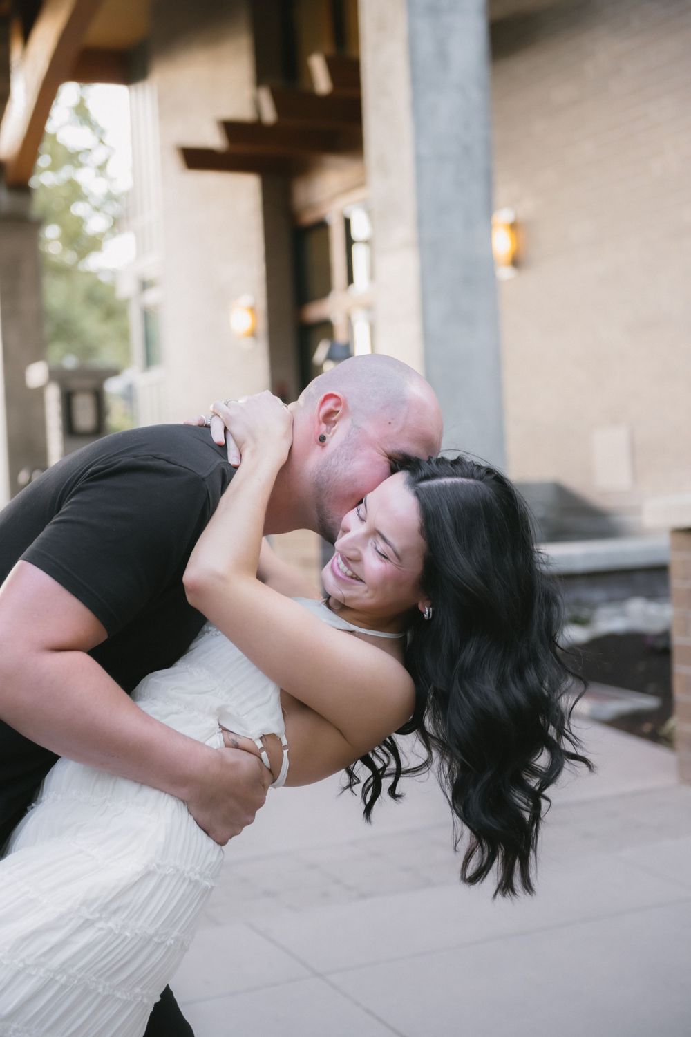 A bald man in a black shirt dips and kisses a smiling woman in a white dress outdoors near a building.