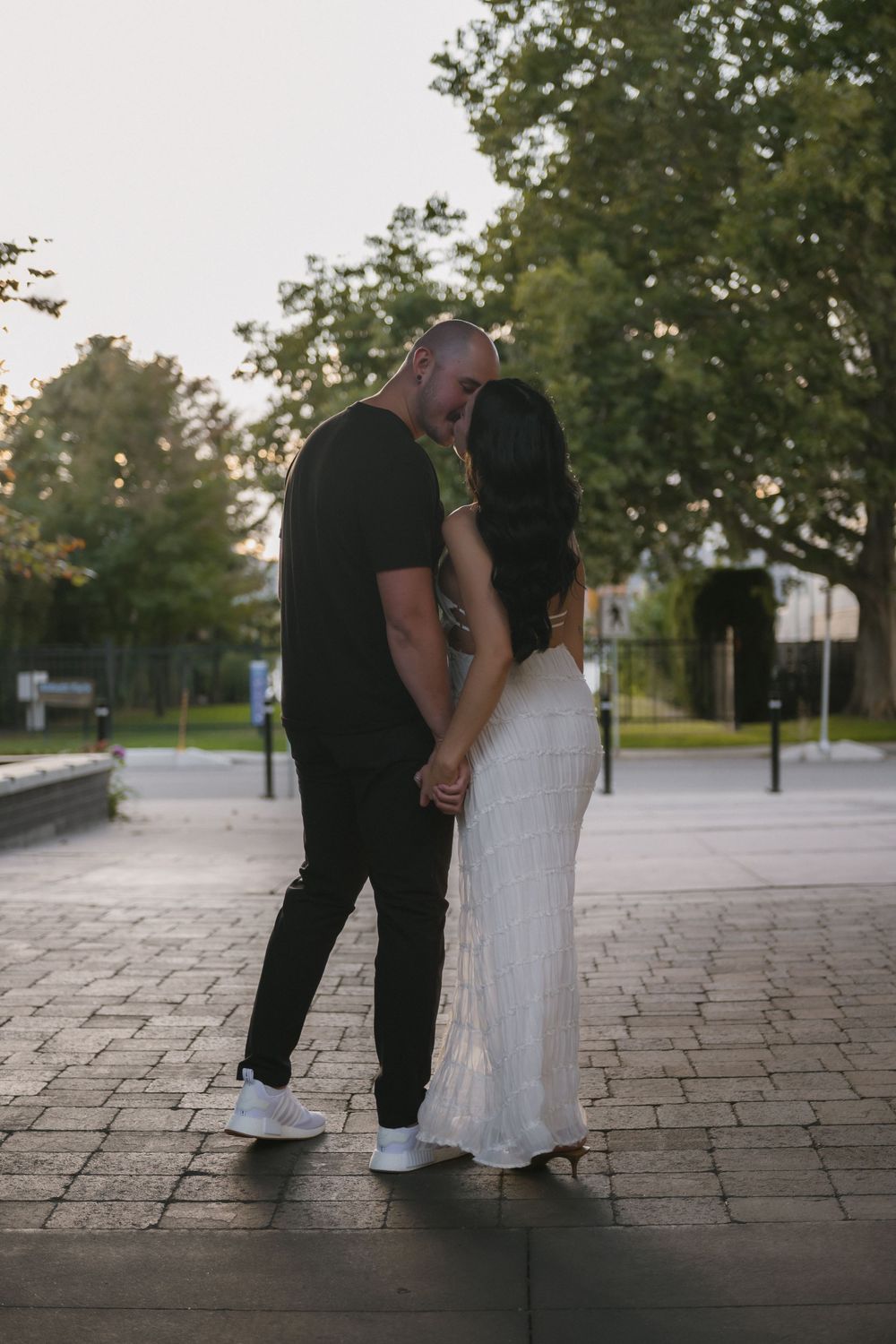 Couple sharing a romantic kiss outdoors on a paved path, man in black outfit, woman in elegant white dress.