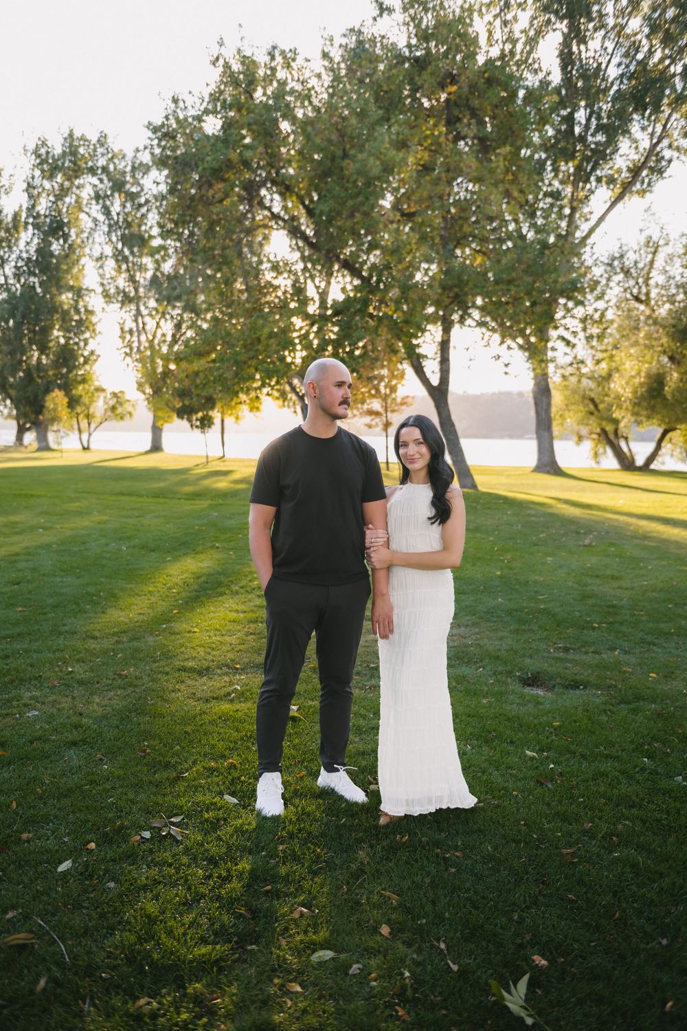 A couple poses in a sunlit park, man in black outfit and woman in white dress, with lush green trees behind them.