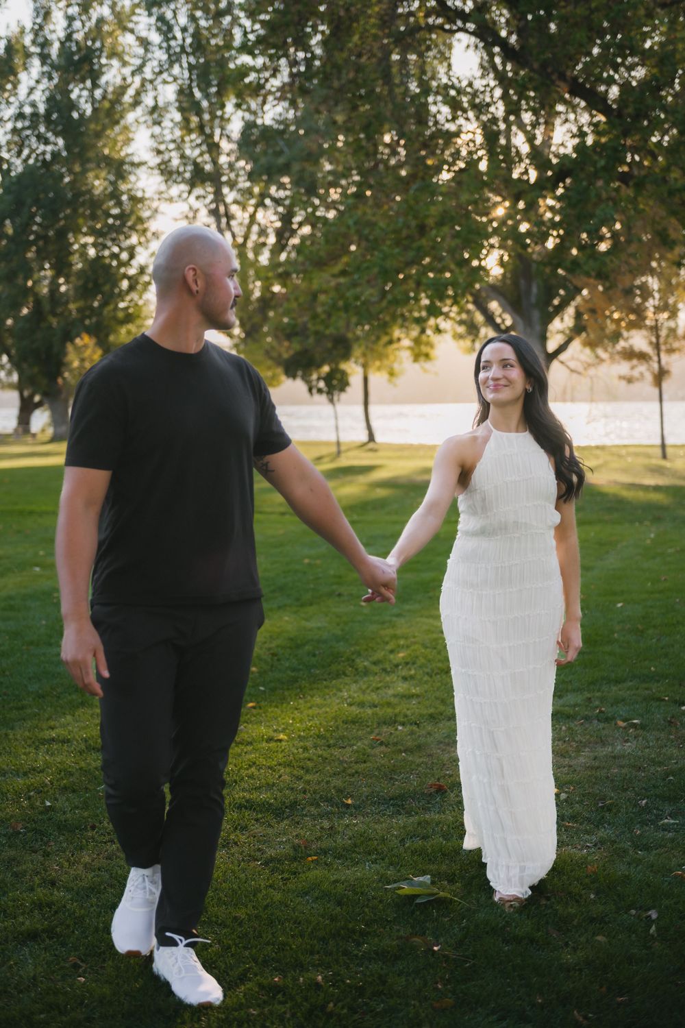 Couple holding hands in a park at sunset, man in black outfit and woman in white dress walking together.