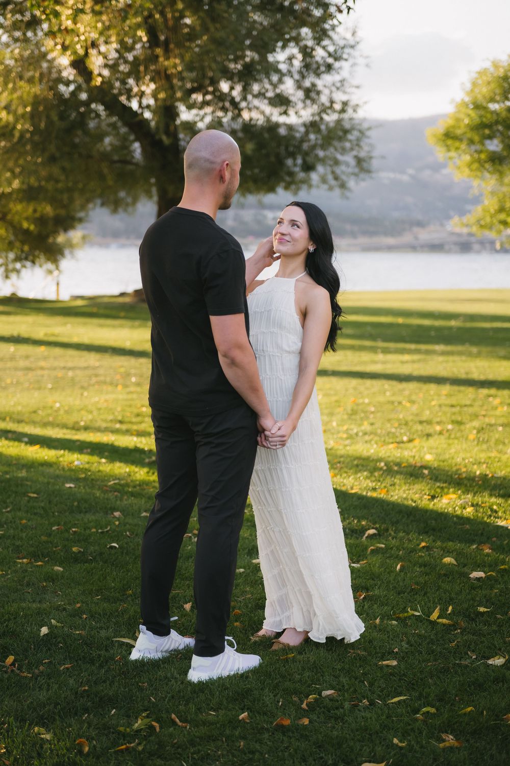 A couple holds hands in a green park by a lake, the woman in a white dress smiling at her bald partner in black.