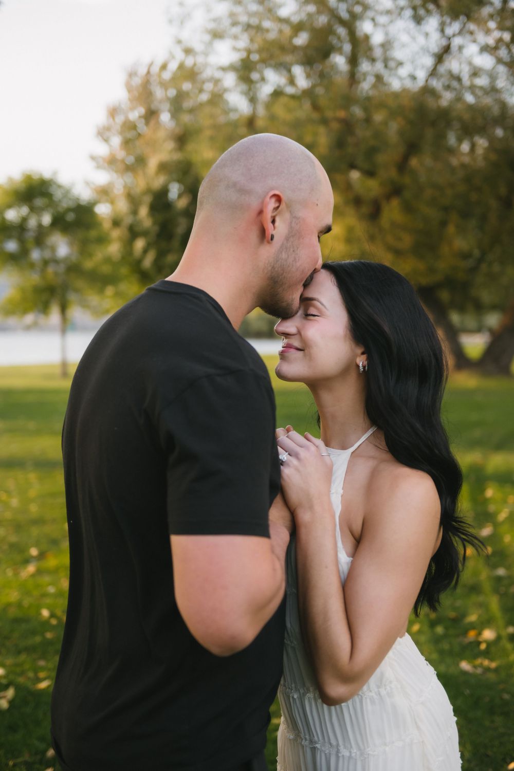 A couple shares a tender forehead touch in a lush green park, the woman smiling in a white dress.