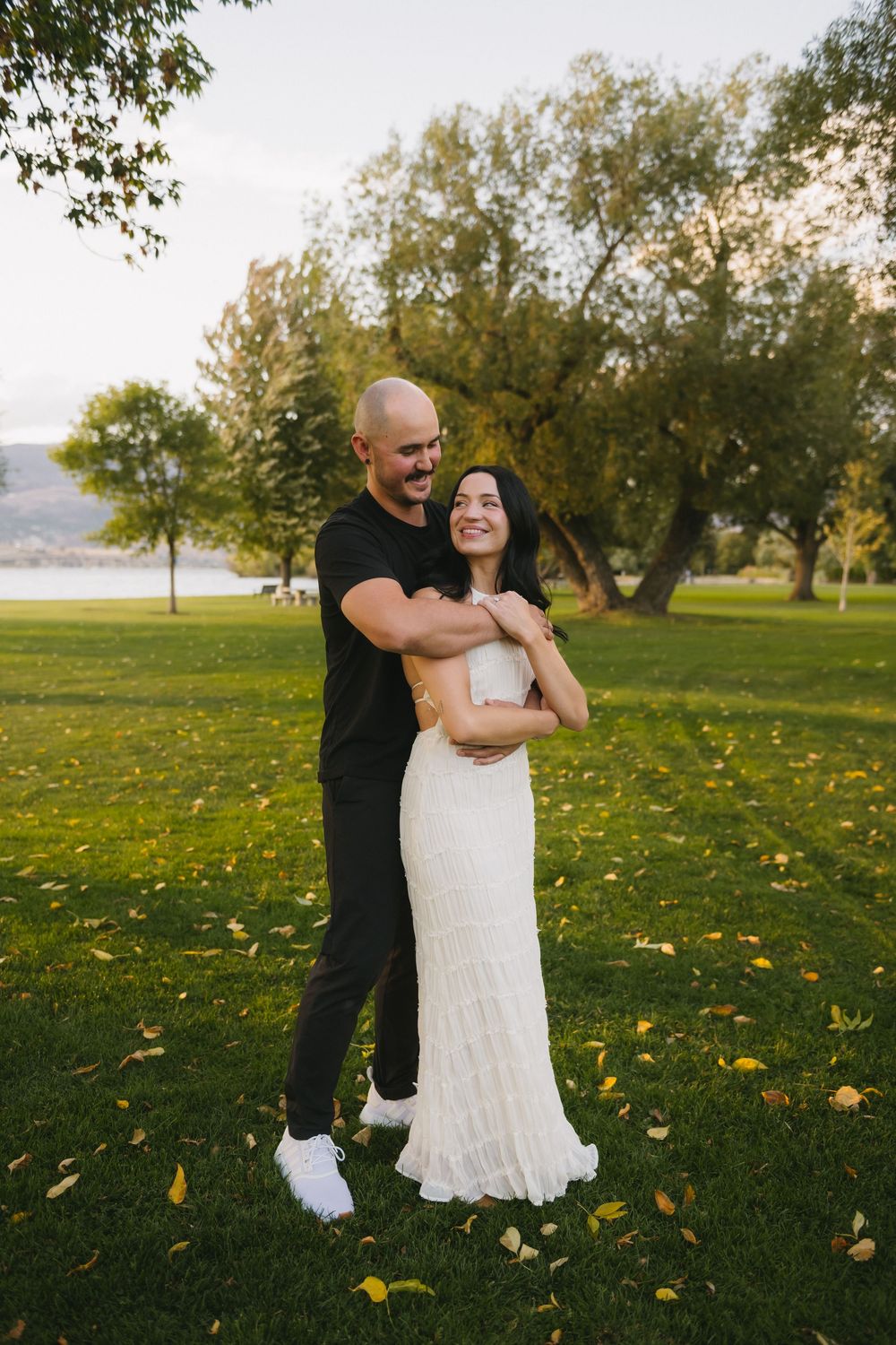 Couple embracing on green grass park, man in black shirt hugging woman in white lace wedding dress, trees background.
