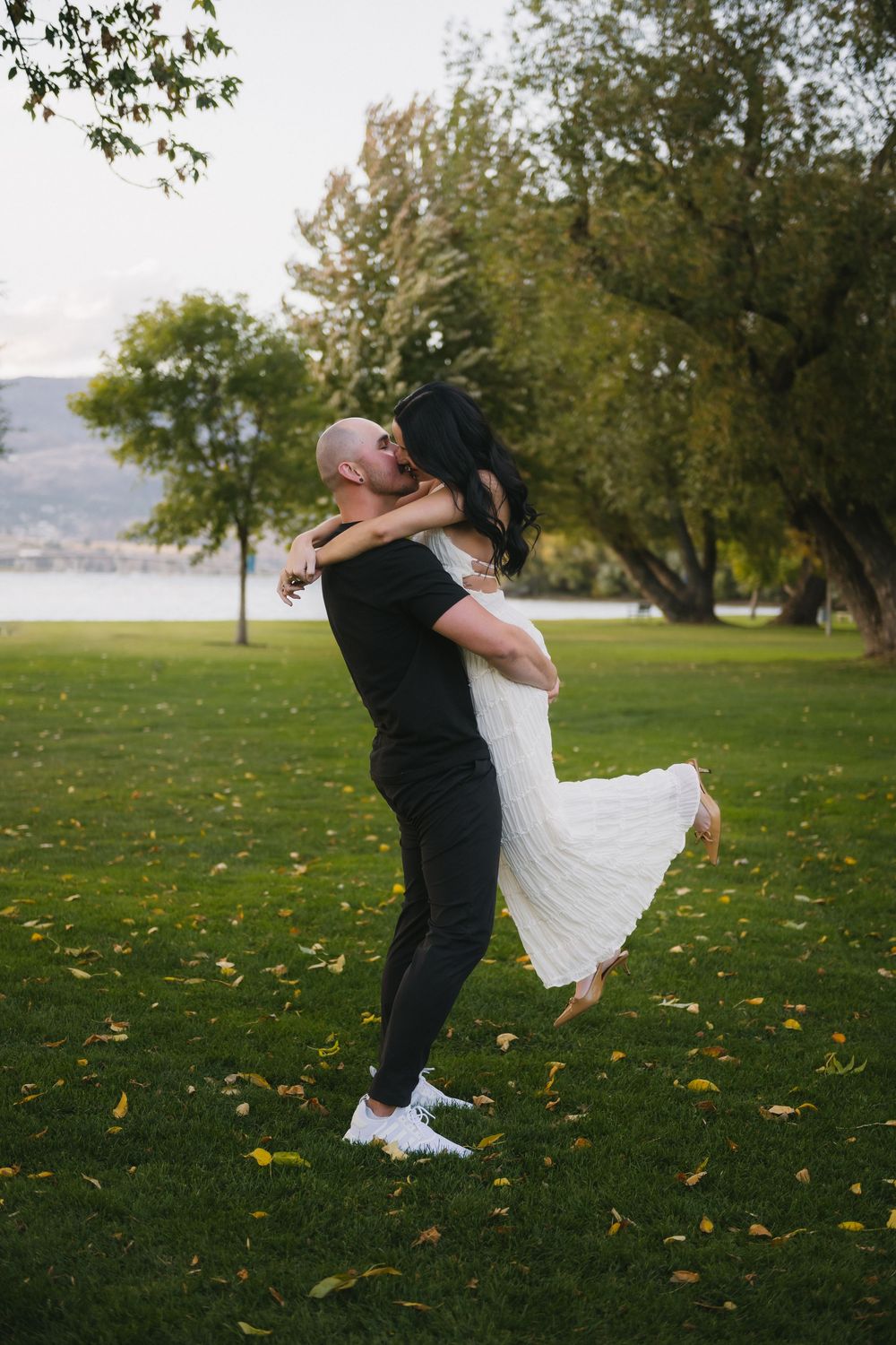 A bald groom lifts his dark-haired bride in a white dress in a lush green park near a lake.