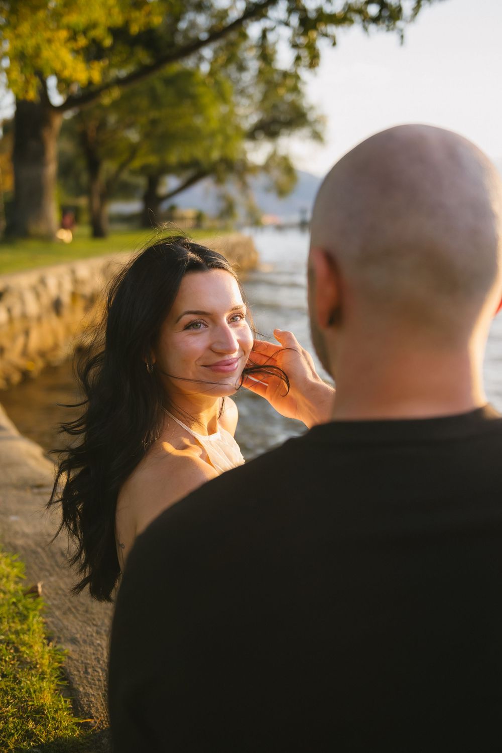 A woman with dark hair smiles warmly at a bald man outdoors near a waterfront during golden hour.