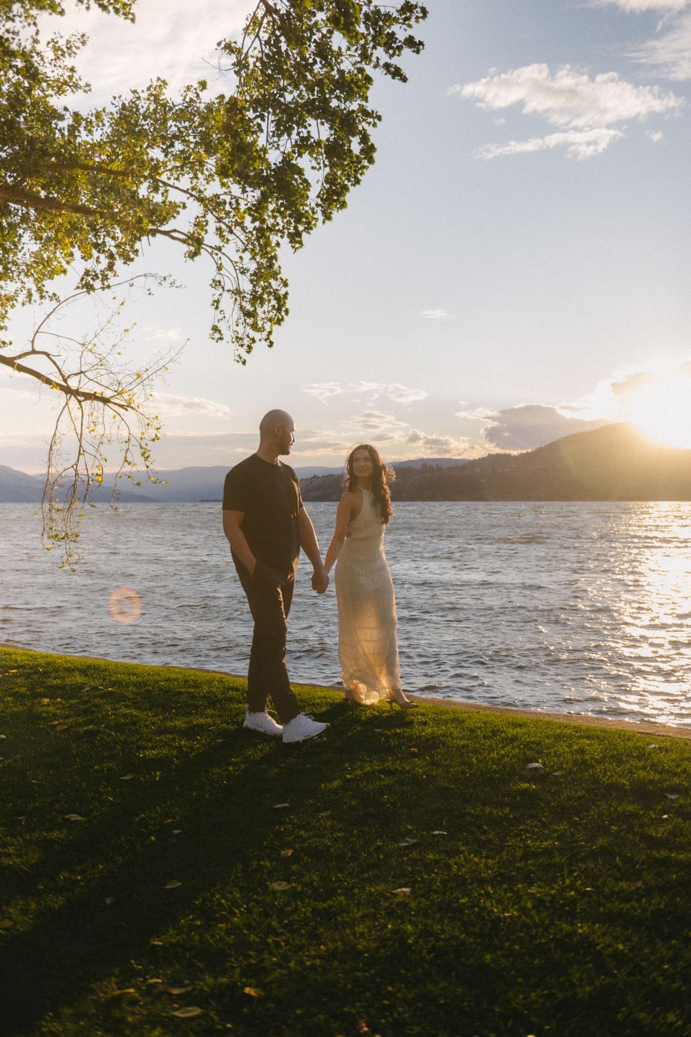 Couple holding hands by a lake at golden hour sunset with mountains in the background.