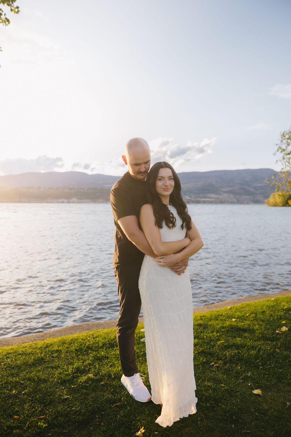 Couple embracing by a lake at golden hour, woman in white dress, man in black outfit, mountains in background.