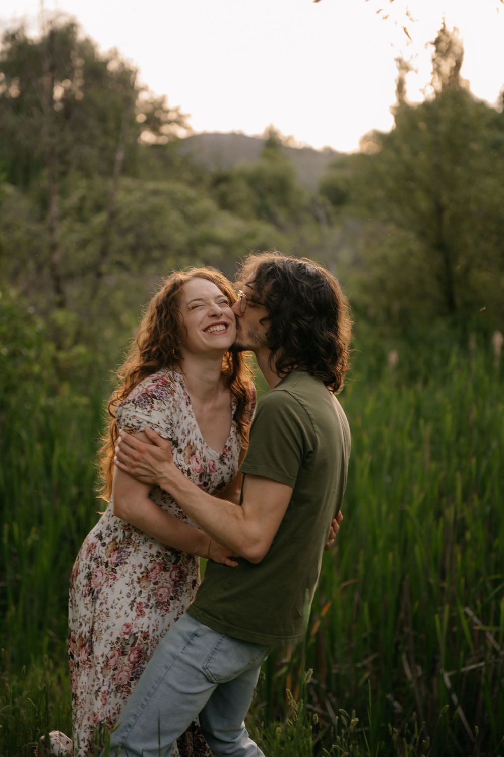 A couple embraces outdoors at sunset, man kissing woman's cheek as she laughs joyfully in a lush green meadow.
