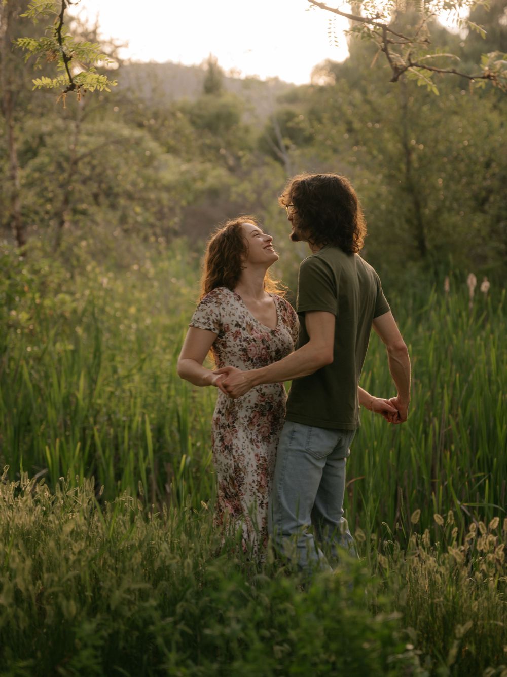 A couple laughs and embraces in a lush green meadow during golden hour sunset light.