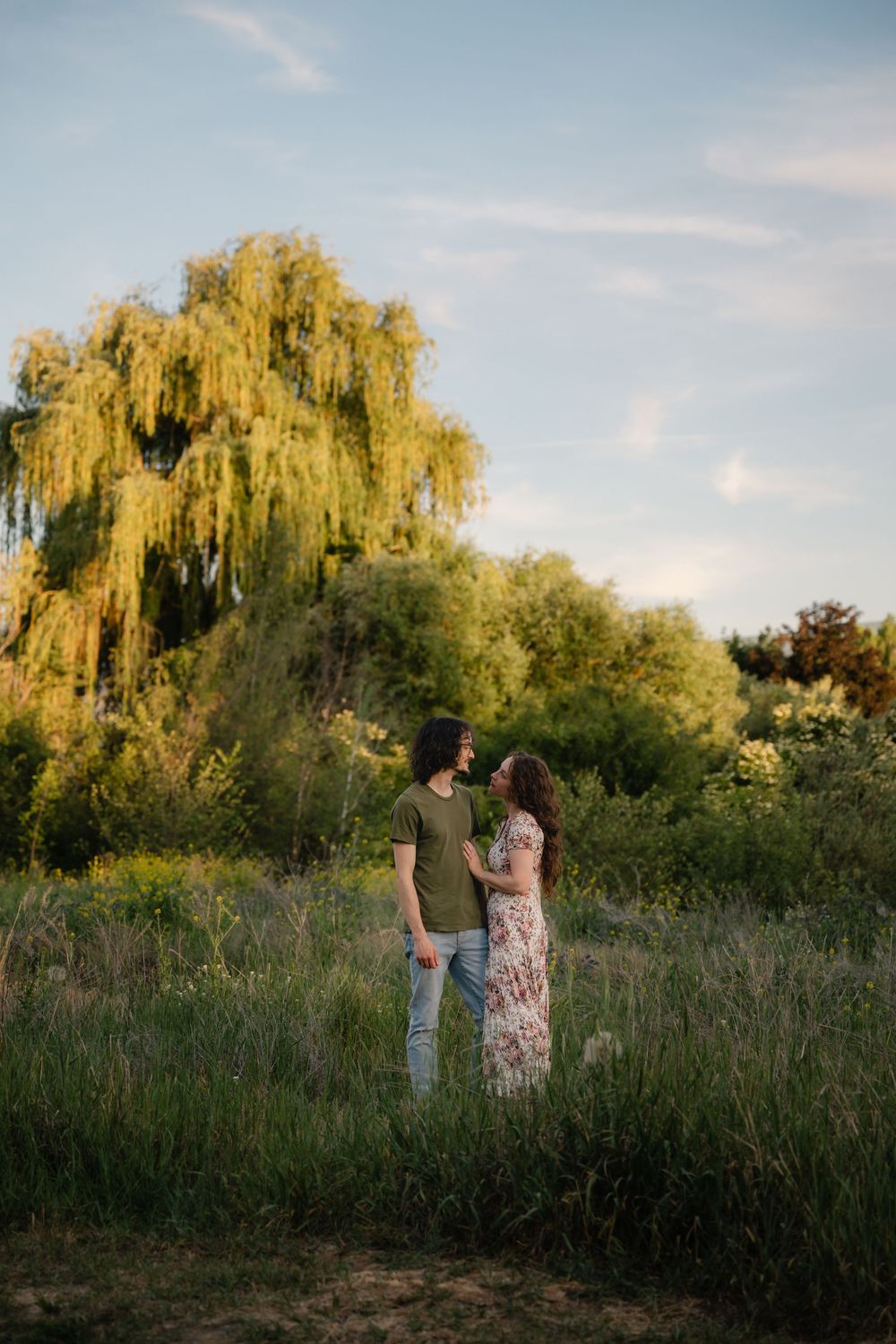 Couple standing together in a lush green field at golden hour, with a weeping willow tree in the background.