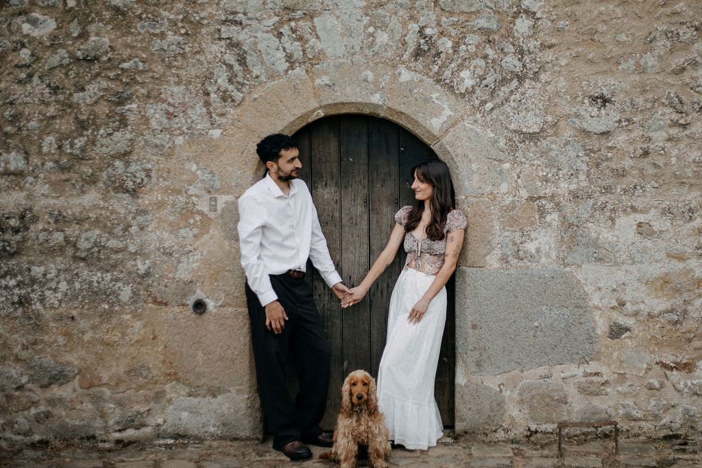 A couple holds hands with their cocker spaniel beside a rustic stone wall and wooden door.