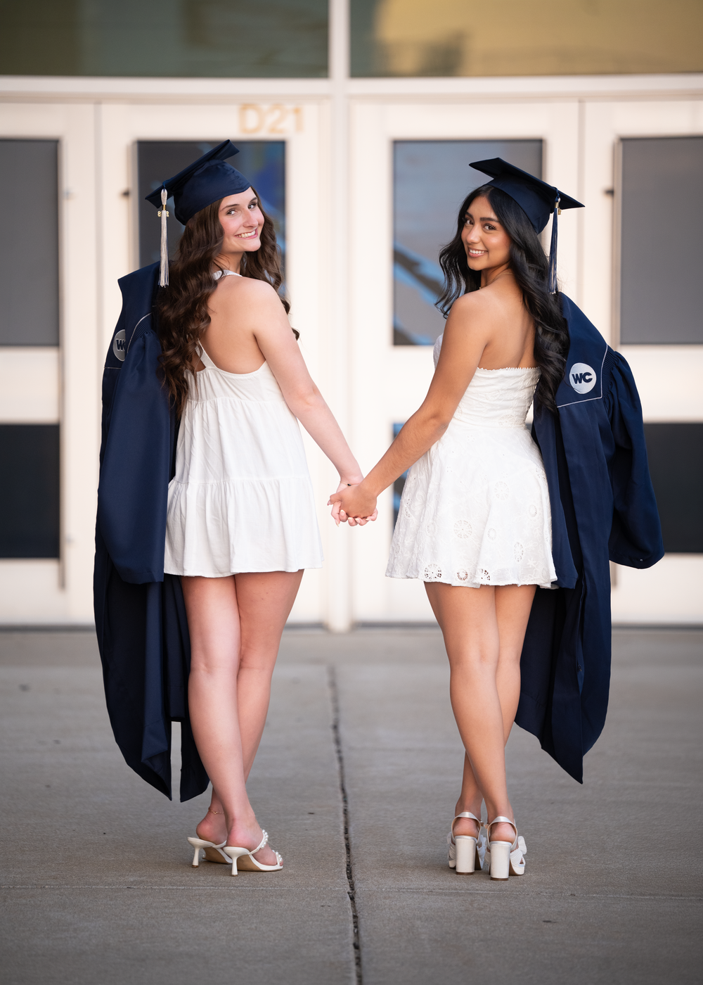 Two female graduates in white dresses and navy caps and gowns hold hands while posing outdoors.