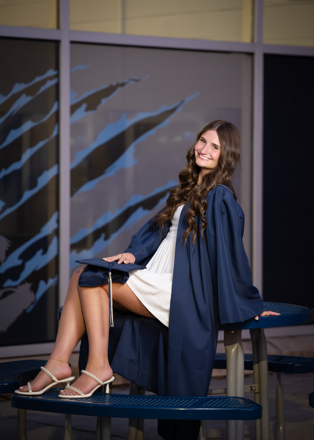 Young woman in navy graduation gown smiles while seated outdoors, wearing a white dress and heeled sandals.