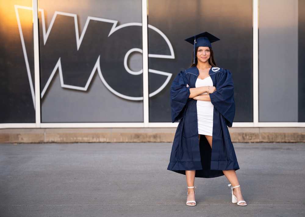 Graduate in navy cap and gown poses confidently in front of a WC building sign, wearing a white dress and heels.