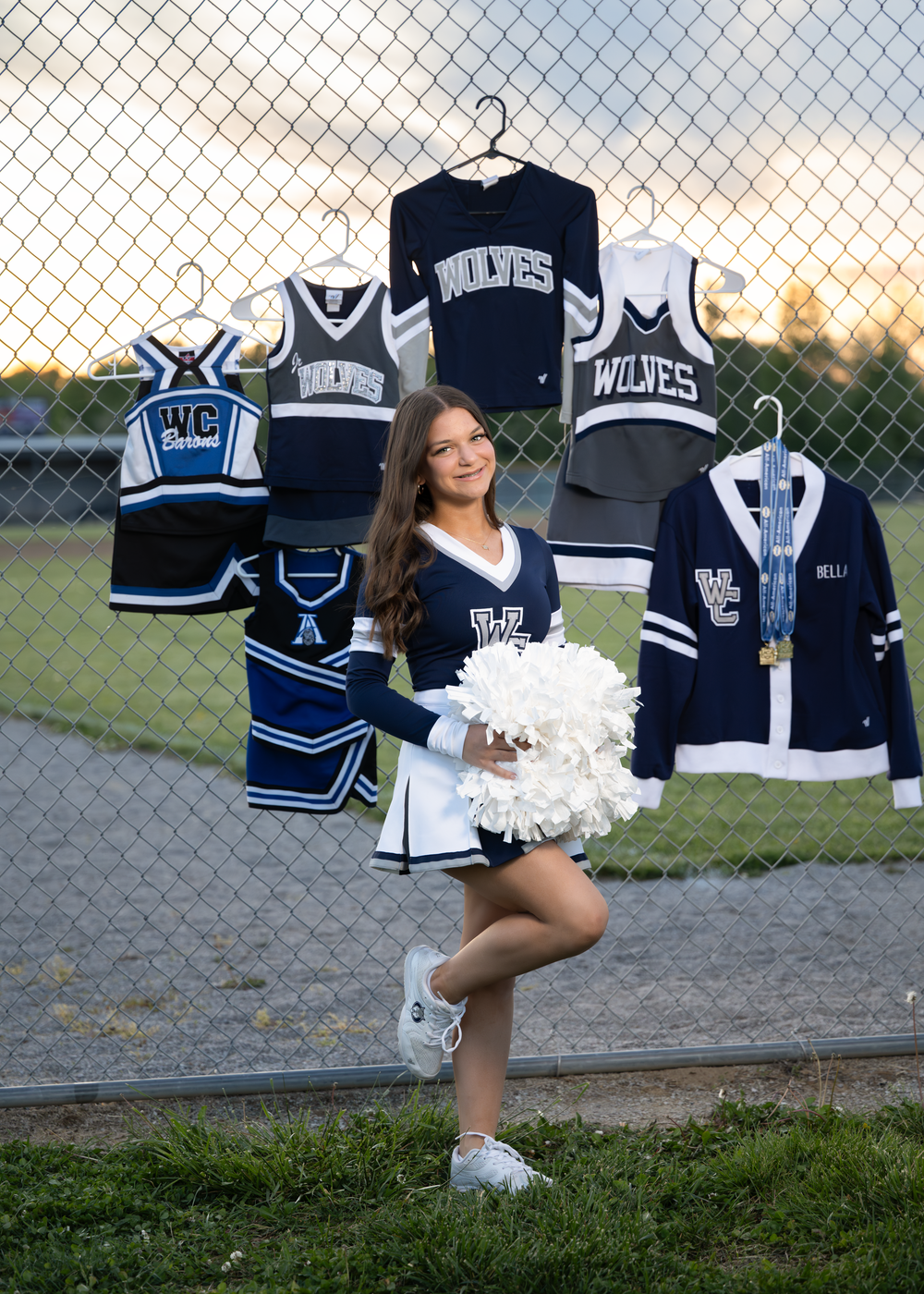 A cheerleader in a Wolves uniform poses with pom-poms in front of a fence displaying her cheer uniforms and letterman jacket.