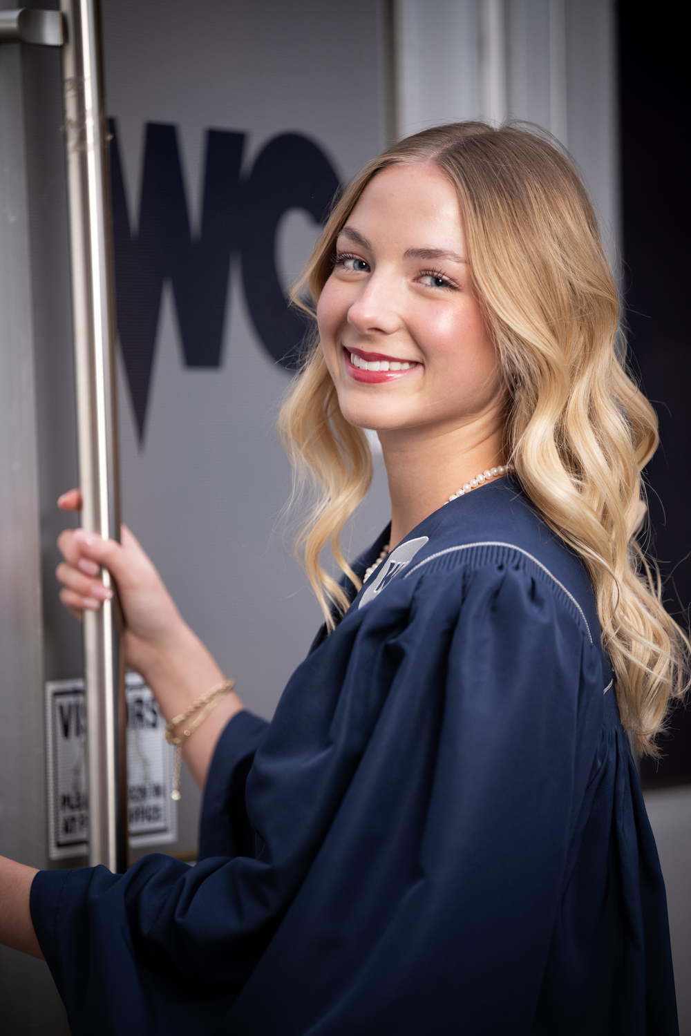 Smiling blonde woman in navy graduation gown holding a metal pole, posing outdoors for a senior portrait photo.