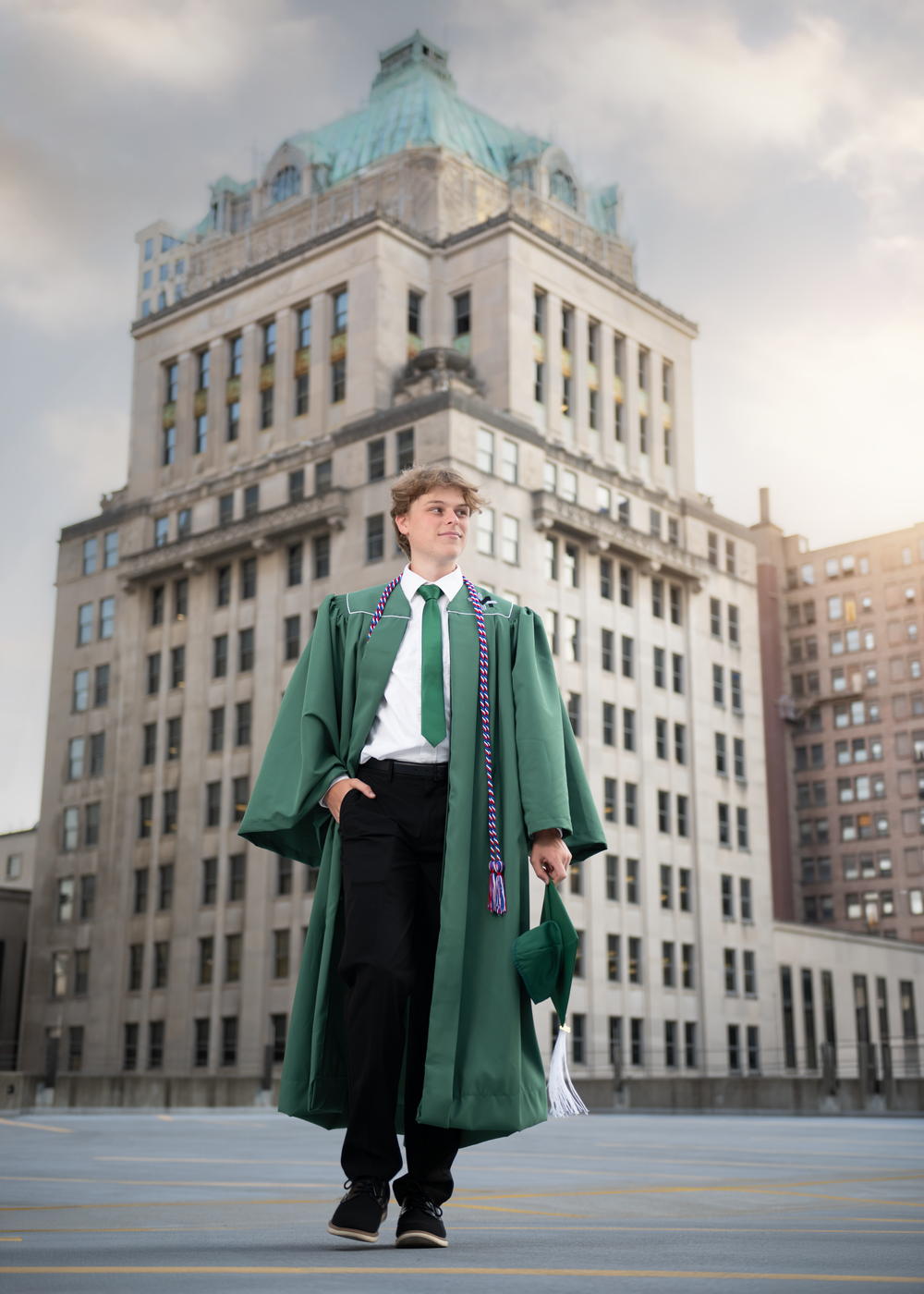 Graduate in green cap and gown stands confidently before a grand stone building in an urban setting.